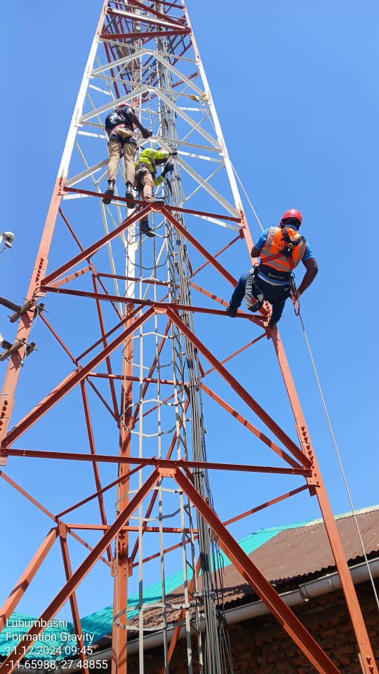 Un groupe d’ingénieurs High-Tech Sarl, équipés de casques de sécurité et de harnais, travaille en hauteur sur une imposante pylône réseau. La scène, baignée par le soleil, capture leur concentration et leur expertise alors qu’ils effectuent des opérations complexes de modernisation des équipements télécoms